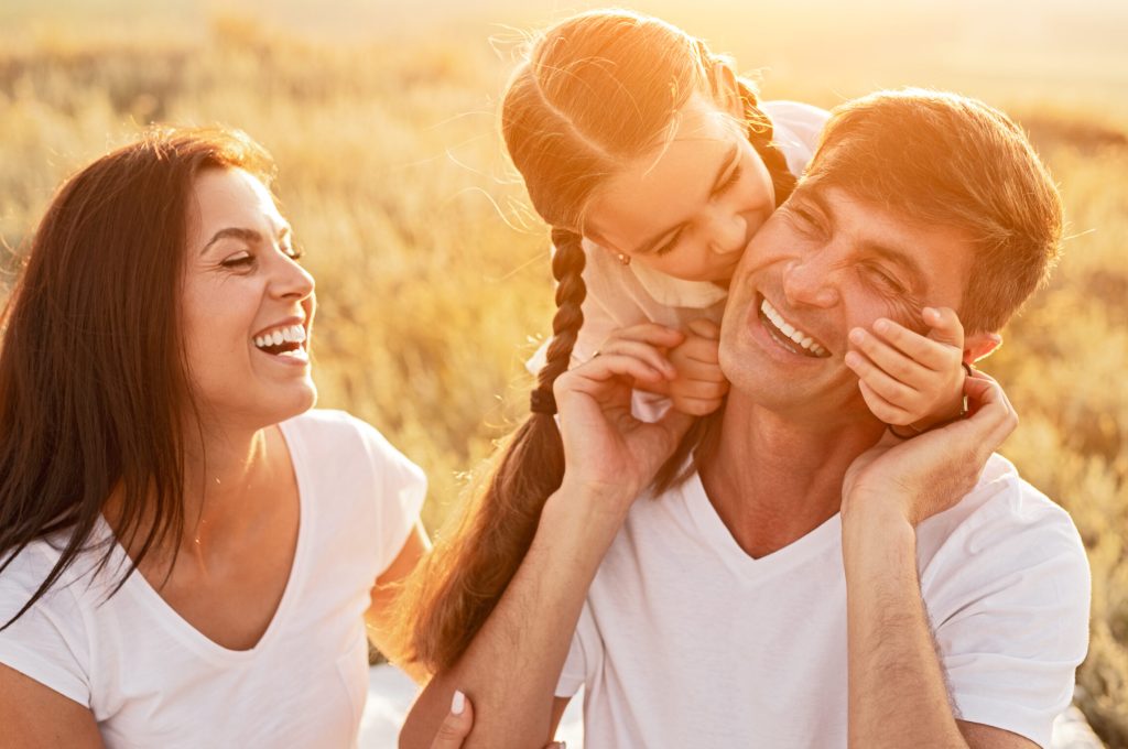 Daughter hugging and kissing cheerful father in cheek near laughing mother while spending day of happy childhood in field